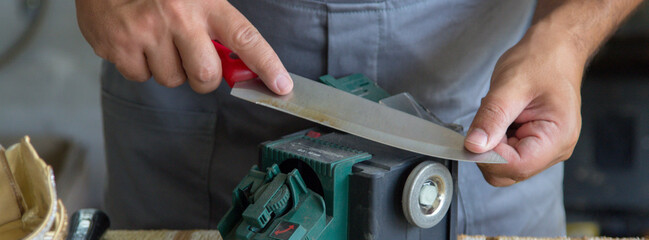 Image of a shoemaker sharpening a kitchen knife with a sharpening wheel. Horizontal banner 