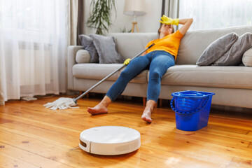 Robotic vacuum cleaner cleaning a room while tired woman relaxing on sofa