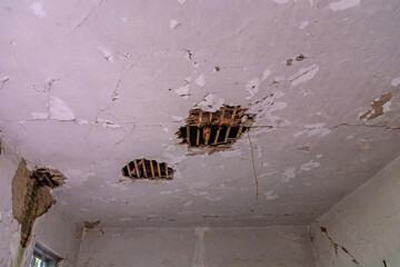 The interior ceiling of an ancient building, riddled with large holes revealing the beams. Cracks run through the ceiling, embodying the passage of time