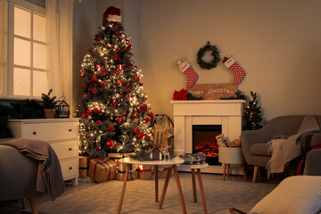 Interior of living room with Christmas tree, fireplace, grey armchairs and chest of drawers at evening