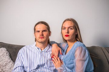An angry couple in light blue clothes, huddled on a sofa, stares ahead