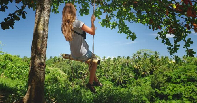 Girl swing on rope sway on the tree in tropical jungle garden. Long hair caucasian woman enjoy outdoor lifestyle travel summer holiday vacation, free happy. Blue sky and green leaf forest. Slow motion