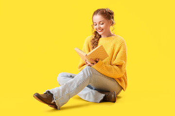 Beautiful happy young woman in warm sweater sitting on yellow background and reading book