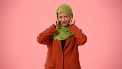 Medium-sized isolated photo capturing an attractive young woman wearing a hijab, veil. She is plugging her ears with fingers and smiling widely.