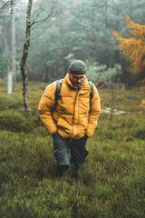 Young man with yellow winter jacket and hat, hands in pockets, walks through birch forest, during cold cloudy autumn day, Decin, Czech republic