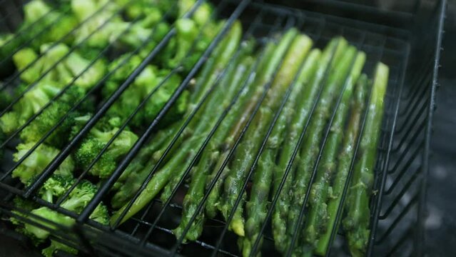 Asparagus and broccoli are being grilled over charcoal on a barbecue outdoors.