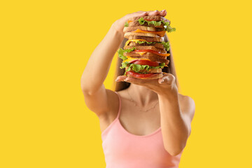 Beautiful young woman holding big sandwich on yellow background