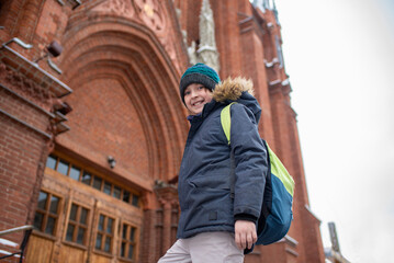 happy child goes to catholic church on sunday in winter. A 10-year-old boy stands in front of a Catholic cathedral, goes to Sunday Christian school. - obrazy, fototapety, plakaty