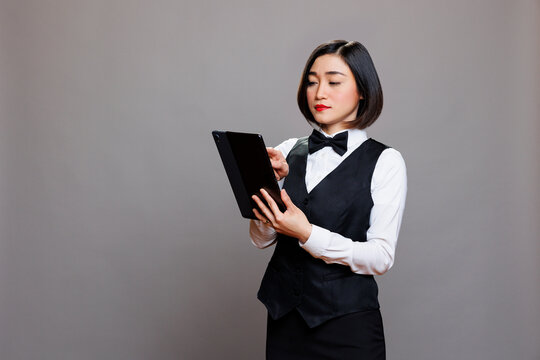 Young asian waitress in uniform checking cafe menu on digital tablet. Catering service staff managing online order and tapping on touchscreen while posing in studio on gray background