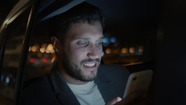 Latin American Man in Modern Car Using Smart Phone. Texting and Smiling. Big City at Night with Traffic Lights and Bokeh. Young Bearded Man using Mobile Phone in Vehicle with Driver.
