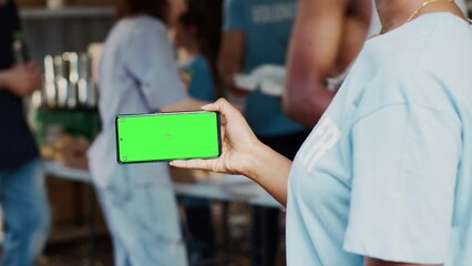 Black woman holds smartphone with isolated chromakey template for customized charitable messages. African American person at an outdoor food drive with a cellphone displaying a green screen.