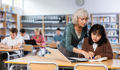 Mature woman teacher working with schoolchildren in the library cheks the assignment from a asian fifteen-old-year schoolgirl - obrazy, fototapety, plakaty