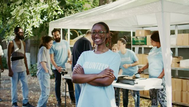 Portrait of african american lady wearing glasses outside with arms crossed and eyes fixed on camera. At food drive, volunteers are aiding the poor, needy and homeless people. Zoom-out, handheld.