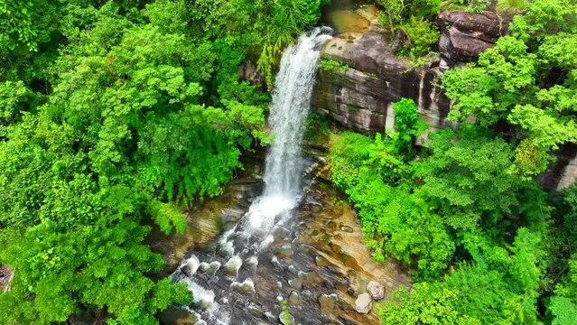 Aerial view of cascading waterfall, lush tropical forest, and towering cliff, captured by a drone. Serene natural beauty. Soi Sawan Waterfall, Pha Taem National Park, Ubon Ratchathani, Thailand.
