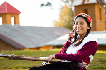 Foto de una feliz campesina caucásica hablando por teléfono inteligente y sosteniendo un tejido de manta artesanía peruana a mano en un atardecer en los Andes peruanos. Mujer usando teléfono móvil - obrazy, fototapety, plakaty