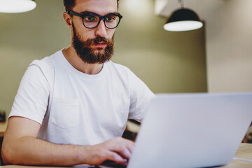 Serious male blogger in spectacles typing text of article for publication on laptop computer