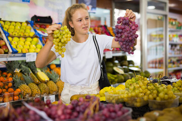 Portrait of a fifteen-year-old girl in a store at the fruit counter, chooses grapes, holding bunches in her hands