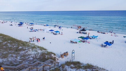 Aerial wooden ram access into Miramar Beach, white sandy shoreline, turquoise water, crowded diverse people swimming, relaxing, colorful tents, canopy, chairs, Destin, Florida