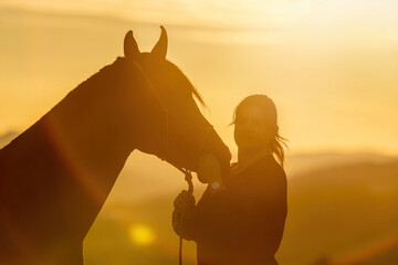 A young female equestrian and her arab berber horsein front of a romantic sunset landscape. Bond...