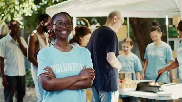 Black lady with spectacles having her arms crossed and gaze fixed on the camera outdoors. Charity workers contributing to non-profit program committed to assisting homeless people. Tripod portrait.