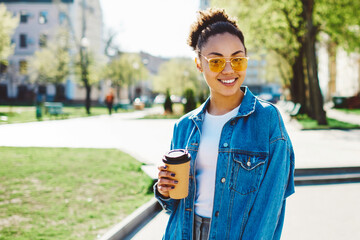 positive woman in trendy apparel looking at camera while holding cup with takeaway coffee