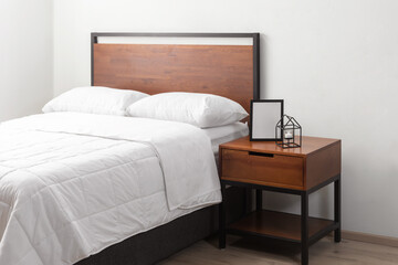 Hotel Bedroom Interior Featuring a King-Size Metal Wood Platform Bed with Headboard, White Cozy Bed Blanket, and a Bedside Table Adorned with a Decorative Ornaments, Set Against a White Wall.
