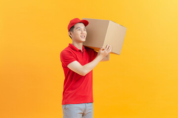 Friendly asian delivery guy wearing red shirt and cap with box parcel on yellow background