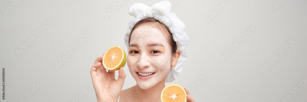 Wall mural Young woman applying clay mask on her mask with sliced fruit in hand over color banner