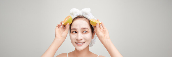 Young woman with facial clay mask holding orange fruit slice covering eyes on white banner
