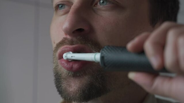 Man brushes his teeth with an electrical tootbrush at morning bathroom before work. Male uses an electric sonic dental cleaning brush. Daily dental hygiene and oral health with eletrical teethbrush. 