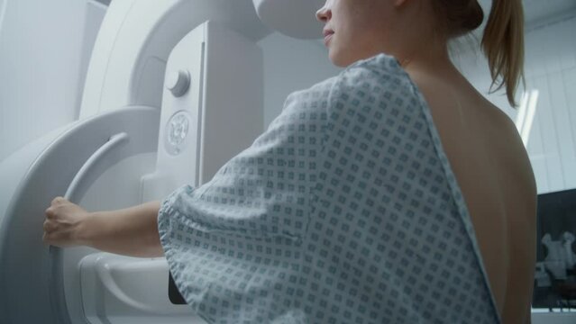 Hospital radiology room. Caucasian adult woman stands during mammography screening diagnostic in clinic. Female doctor finishes procedure, turns off modern mammogram machine. Breast cancer prevention.