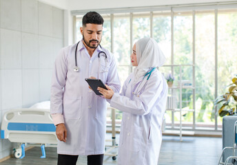 Asian Indian professional successful bearded male doctor in white lab coat uniform with stethoscope standing talking discuss brainstorm with female muslim colleague in hijab holding tablet computer