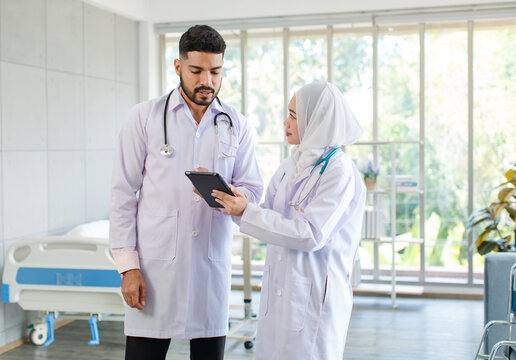 Asian Indian professional successful bearded male doctor in white lab coat uniform with stethoscope standing talking discuss brainstorm with female muslim colleague in hijab holding tablet computer