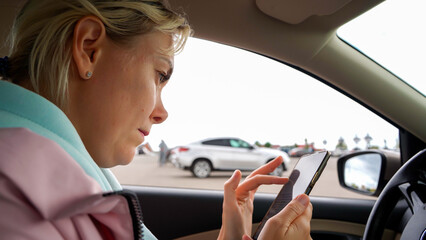 Naklejka premium A female driver uses a mobile phone while driving a car. Attractive young female driver uses smartphone to navigate or call emergency help. A young natural beauty woman uses a phone during a road trip