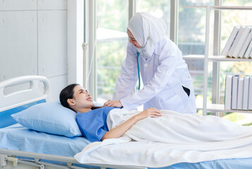 Obraz premium Asian professional successful experienced muslim female doctor in white lab coat with hijab using stethoscope listening heartbeat of healthy patient in blue uniform laying on bed in hospital ward
