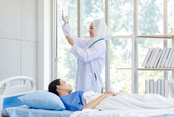 Asian professional successful experienced muslim female doctor in white lab coat with hijab using stethoscope listening heartbeat of healthy patient in blue uniform laying on bed in hospital ward