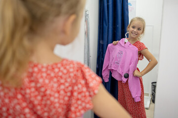 A cute little girl chooses new clothes for herself in the store. Tries on things in the fitting...