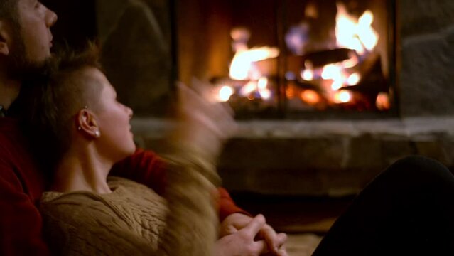 bearded man and handsome non binary girl with  short haircut and piercings are relaxing in the evening near the fireplace. Drinking tea in the cozy atmosphere of a hunting lodge, sitting near the fire