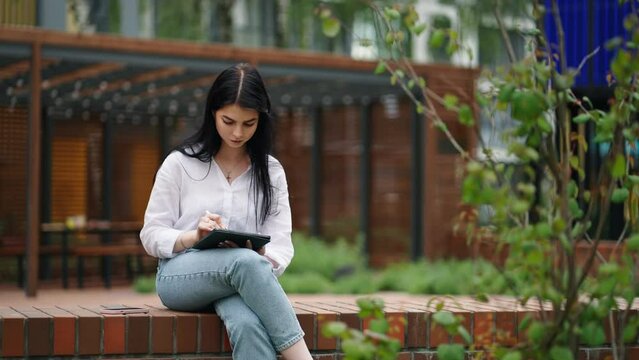 Young Adult Woman Resting In Park In Daytime And Using Graphics Tablet With Stylus For Work