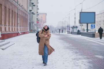 A girl in a long coat walks through the city in cold weather - obrazy, fototapety, plakaty