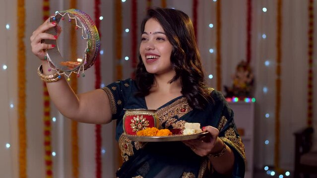 Newly-wed Indian woman celebrating Karwa Chauth festival in traditional wear - wearing traditional saree . Beautiful wife happily looking at the moon and then at her husband through a sieve - Karwa...