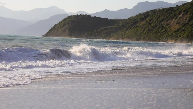 Waves crushing on a beach in slow motion with cliffside and mountains on the background