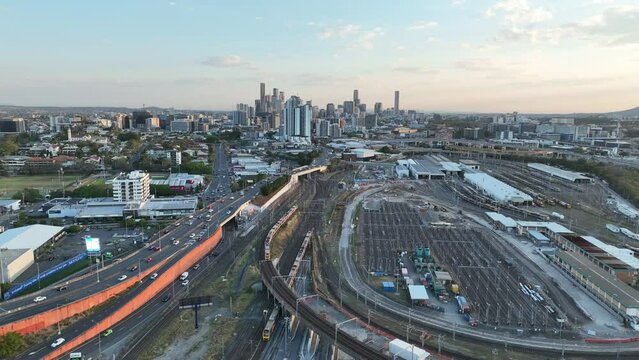 Drone shot tracking train crossing Brisbane City Mayne Railway yard as camera pulls away revealing trains driving below. City Skyline standing tall against lovely sunset sky.