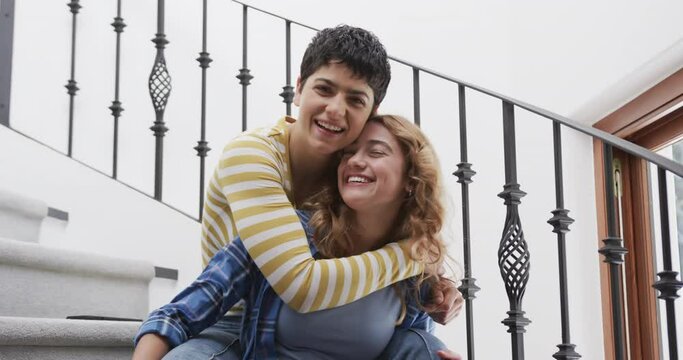 Happy caucasian lesbian couple sitting on stairs, embracing and smiling in sunny house