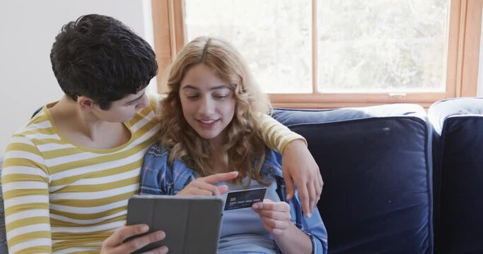 Happy caucasian lesbian couple embracing on sofa, using tablet and credit card in sunny house