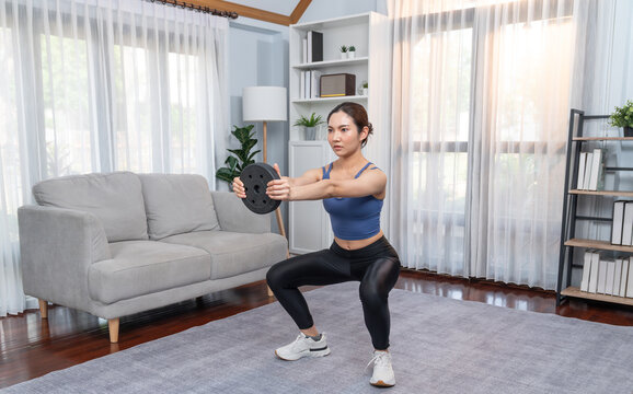Vigorous energetic woman doing squat weight lifting exercise at home. Young athletic asian woman strength and endurance training session as home workout routine with squat.
