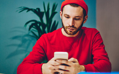 Serious bearded guy in red hat checking mail on smartphone while sitting in coworking office