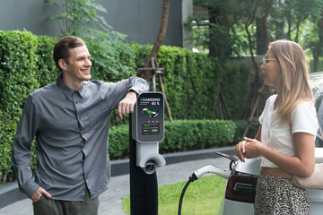 Young couple travel with EV electric car charging in green sustainable city outdoor garden in...