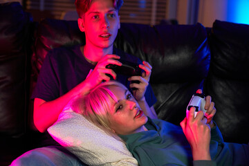 Young caucasian couple playing video game with game pads while sitting and lying on sofa