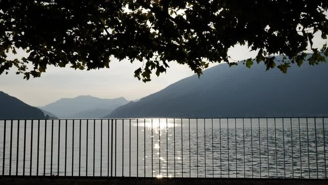 Waterfront with Railing and Tree Branch over Lake Lugano with Mountain in Sunset in Bissone, Ticino, Switzerland.